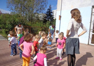 Dzieci z najmłodszej grupy stoją w swobodnej grupie na tarasie. Przed nimi stoi nauczycielka i trzyma ręce na biodrach. Dzieci próbują ją naśladować.